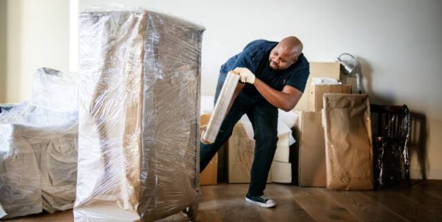 Man wrapping a tall piece of furniture with plastic wrap in a room full of packed boxes and packing materials.